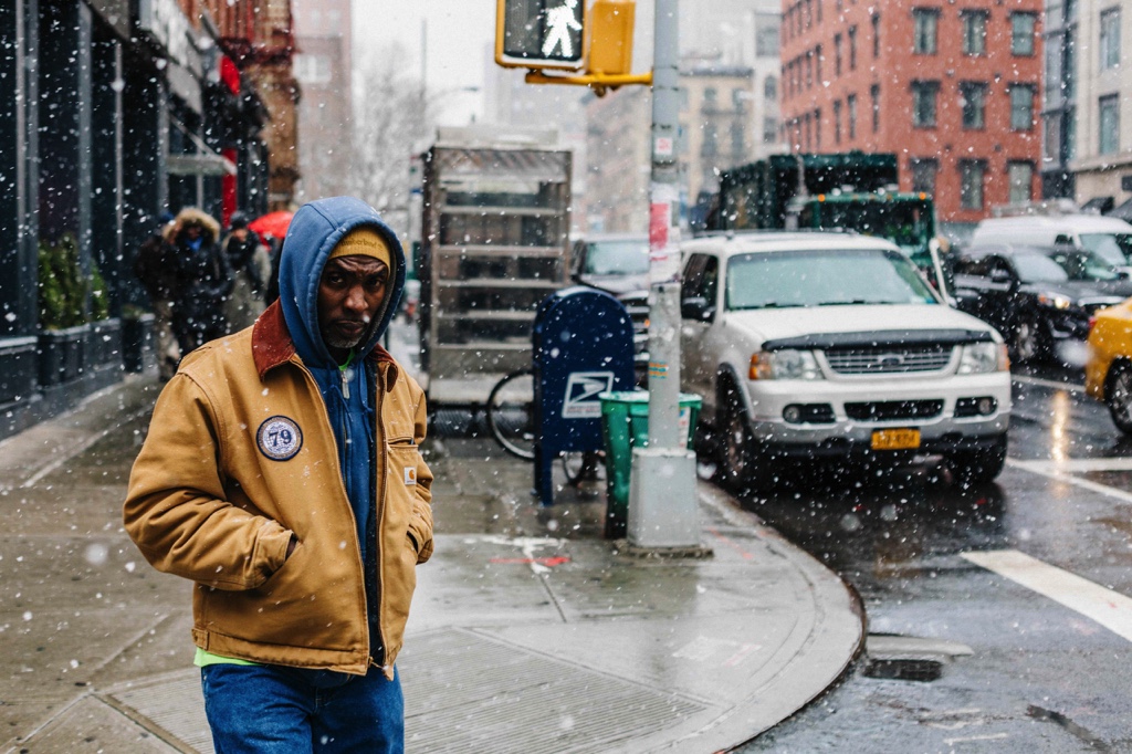 A man crossing the street on a snowy day in Manhattan, New York. He is looking straight at the camera.