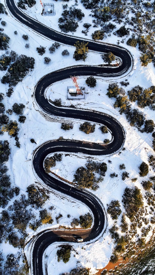 Winding black road through a snowy landscape with some trees, viewed from above.