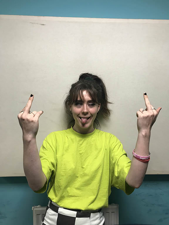 Colour photograph of a woman wearing a yellow top, sticking up two fingers.